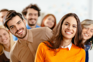 A group of cheerful, diverse young professionals standing in a diagonal line, leaning playfully toward the camera and smiling. The setting appears to be a bright, modern office space, suggesting a positive and collaborative team culture.